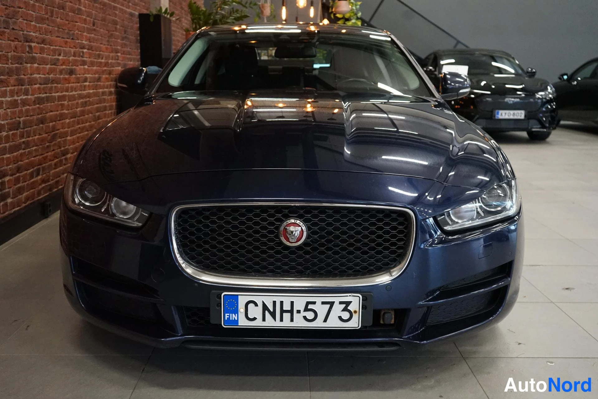 Edestä: Etunäky tummansinisestä autosta: kuusikulmainen jäähdytinsäleikkö Jaguar-merkillä ja rekisterikilpi.
