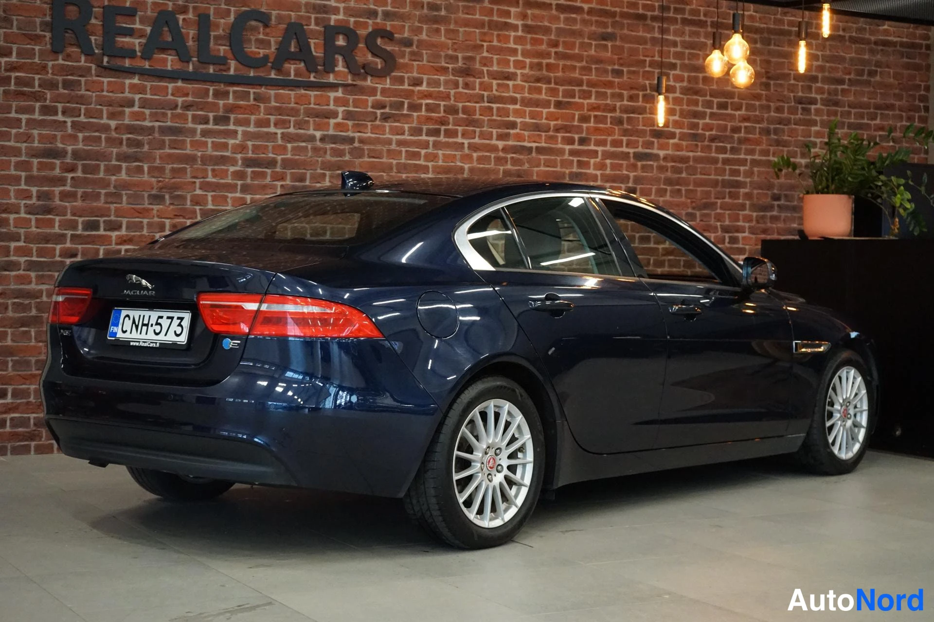 Takaviisto: Tummasininen Jaguar XE -sedan takaa sivulta kuvattuna autotallin tiiliseinää vasten, rekisteritunnus.