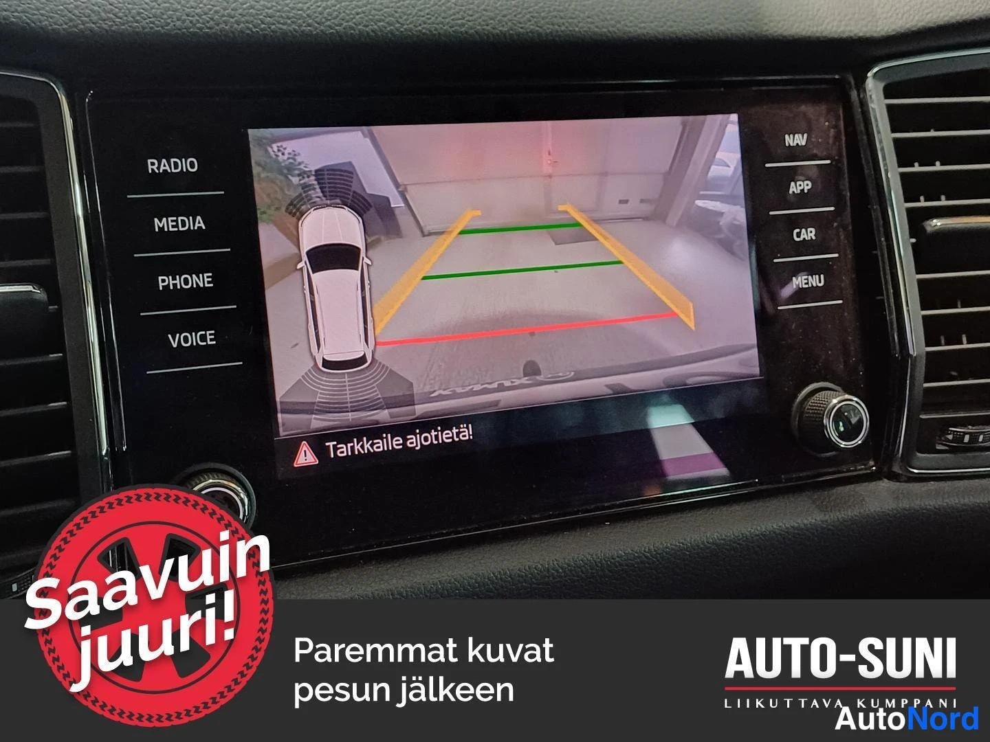 Yksityiskohta: Infotainment-näyttö näyttää peruutuskameran kuvan ohjausviivoilla, tekstillä Tarkkaile ajotietä!, sekä sivuvalikot RADIO, MEDIA, PHONE, VOICE ja NAV, APP, CAR,