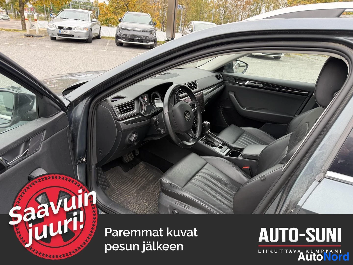 Muu: Avatun etuoven takaa näkyvä auton sisätilä: ohjauspyörä, kosketusnäyttö, automaattivalitsin ja mustat nahkaistuimet.