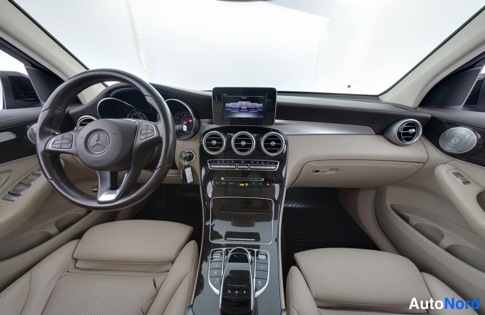 Sivulta: Mercedes-Benzin ohjaamo etupäästä: beige nelipaikkainen sisusta, ohjauspyörä, kolme pyöreää ilmastointiventtiiliä ja kosketusnäyttö keskikonsolissa.