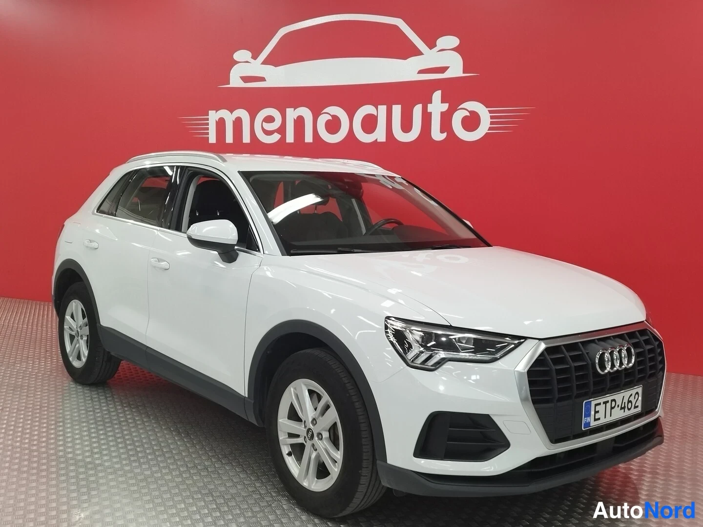 Etuviisto, nokka oikealle: Valkoinen Audi Q3 -maastoauto kolmannelta etunäkymältä oikealta, etusäleikkö ja rekisterikilpi näkyvissä punaista taustaa vasten.