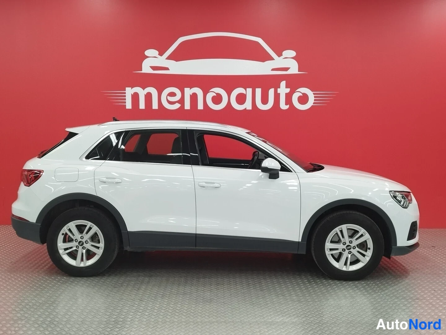 Etuviisto, nokka oikealle: Valkoinen Audi Q3 -maastoauto sivulta vasemmalta nähtynä, molemmat sivut ja pyörät näkyvissä punaista mainostaulua vasten.