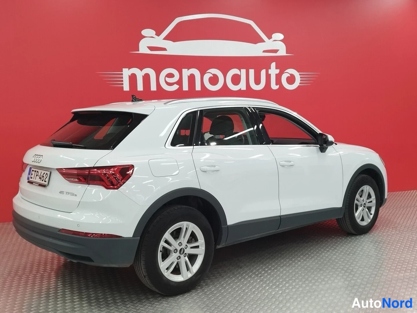 Takaviisto: Valkoinen Audi Q3 -maastoauto takaosan ja vasemman sivun näkymässä, takavalot ja rekisterikilpi näkyvissä punaista taustaa vasten.