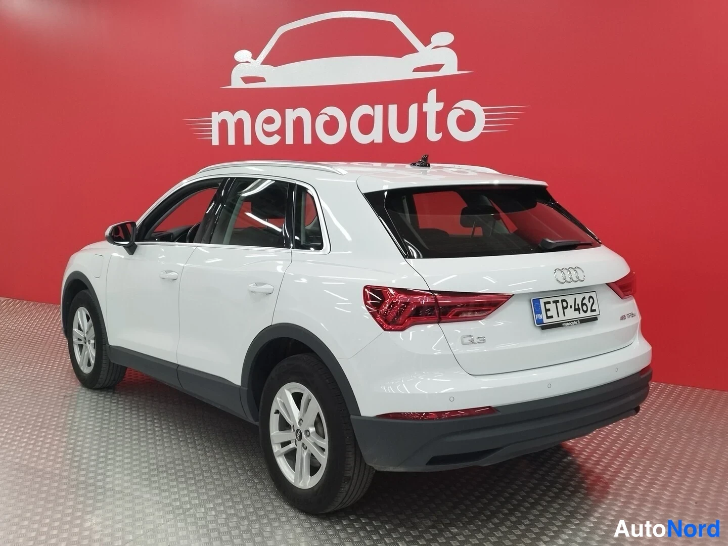 Takaviisto: Valkoisen Audi Q3:n takakulmanäkymä vasemmalta, rekisterikilpi, punainen menoauto-taustaseinä.