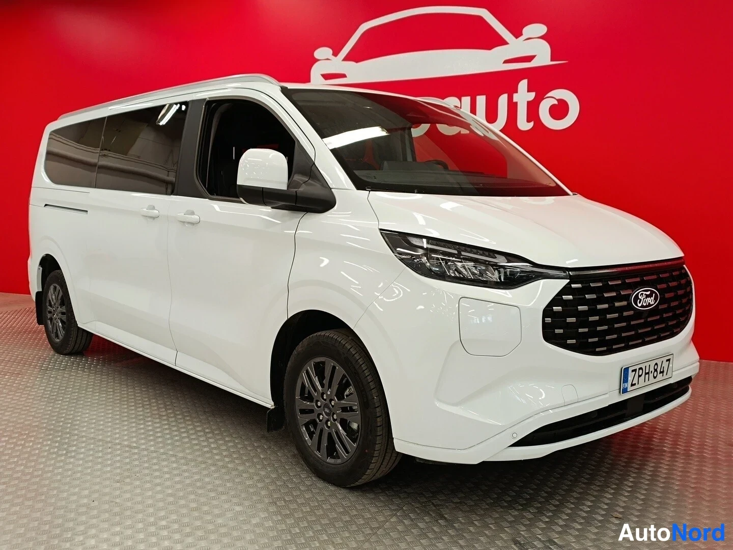 Etuviisto, nokka oikealle: Valkoinen Ford Tourneo Custom -pakettiauto kuvattu kolmanneskulmassa edestä oikealta punaisen taustan edessä; kylki ja etusäleikkö näkyvät, rekisteritunn
