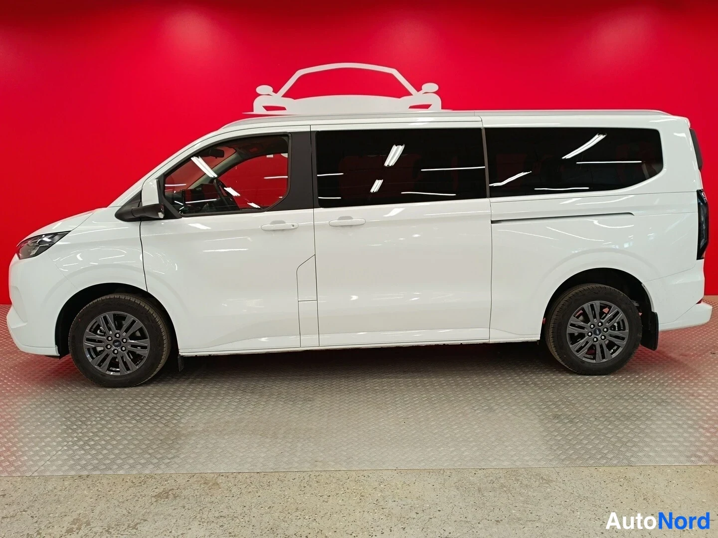 Etuviisto, nokka vasemmalle: Valkoinen Ford Tourneo Custom -pakettiauto kuvattu vasemmalta sivulta punaisen taustan edessä; pitkä matkustamoyksikkö, tummat ikkunat ja kevytmetalliv