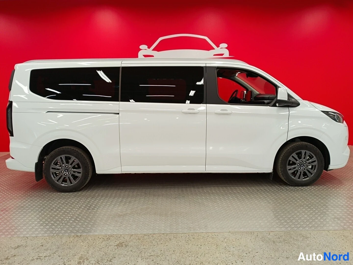 Takaviisto: Valkoinen Ford Tourneo Custom -pakettiauto kuvattu takaviistosta vasemmalta punaisen taustan edessä; vasen kylki ja takavalojen reuna näkyvät, kevytmetallivante