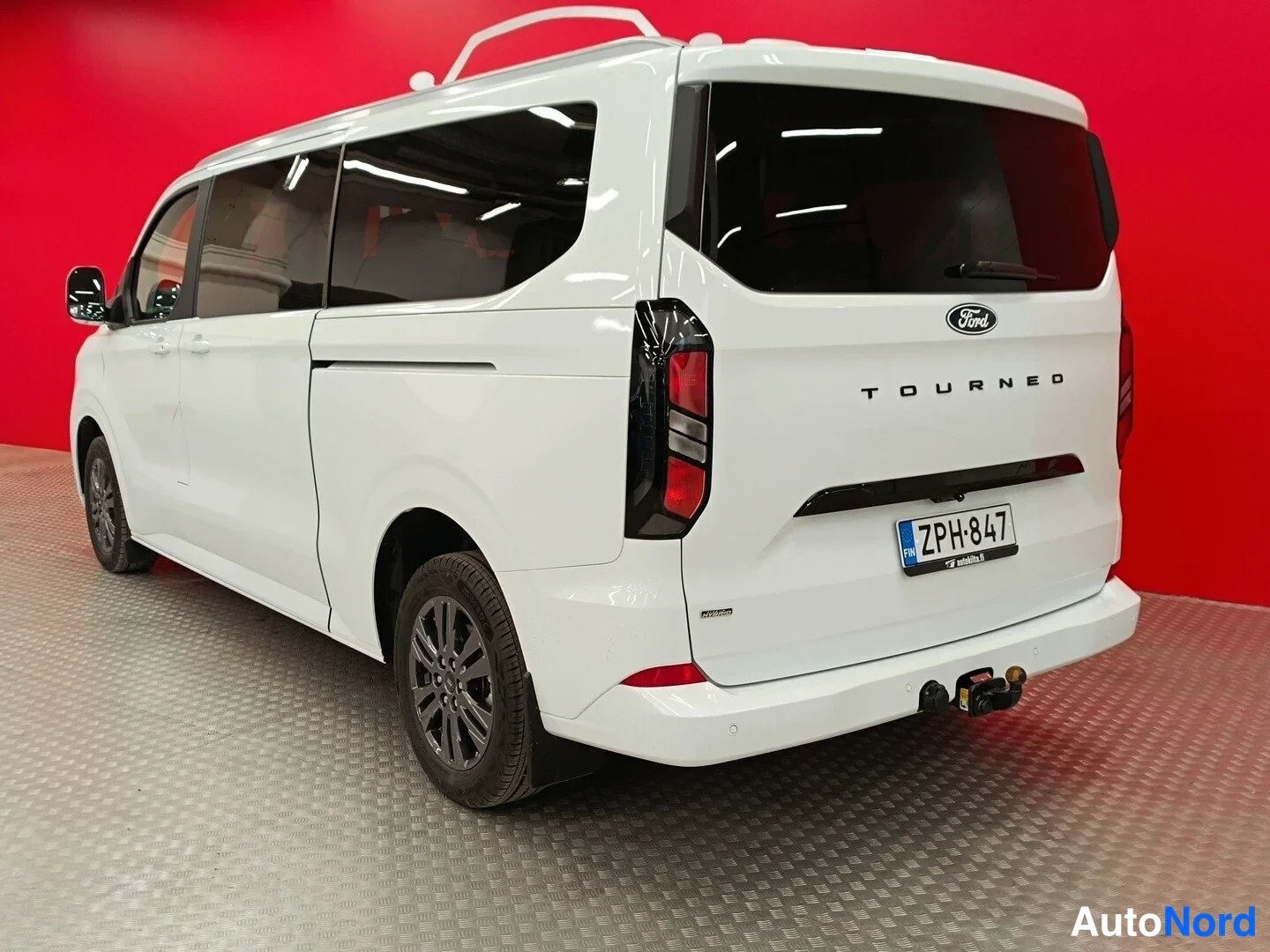 Takaviisto: Valkoisen Ford Tourneo -pakettiauton takaosa ja vasen sivu punaisella taustalla; rekisterikilpi ja takahana näkyvissä.