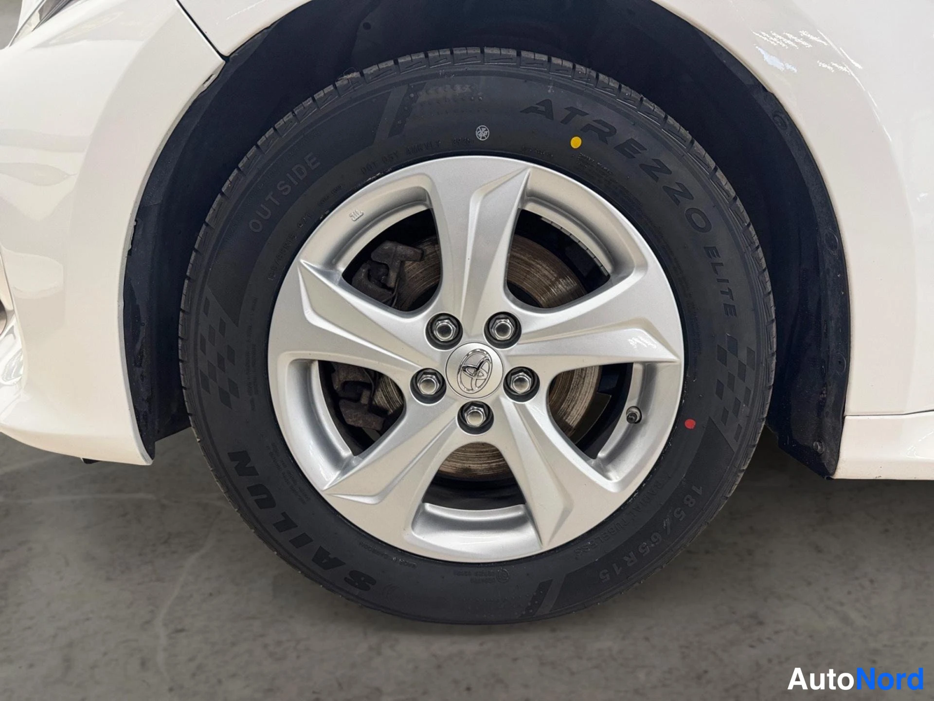 Sivulta: Valkoisen auton etupyörän lähikuva: hopeinen viisipuomainen Toyota-vanne ja Sailun Atrezzo Elite -rengas 195/65 R15.