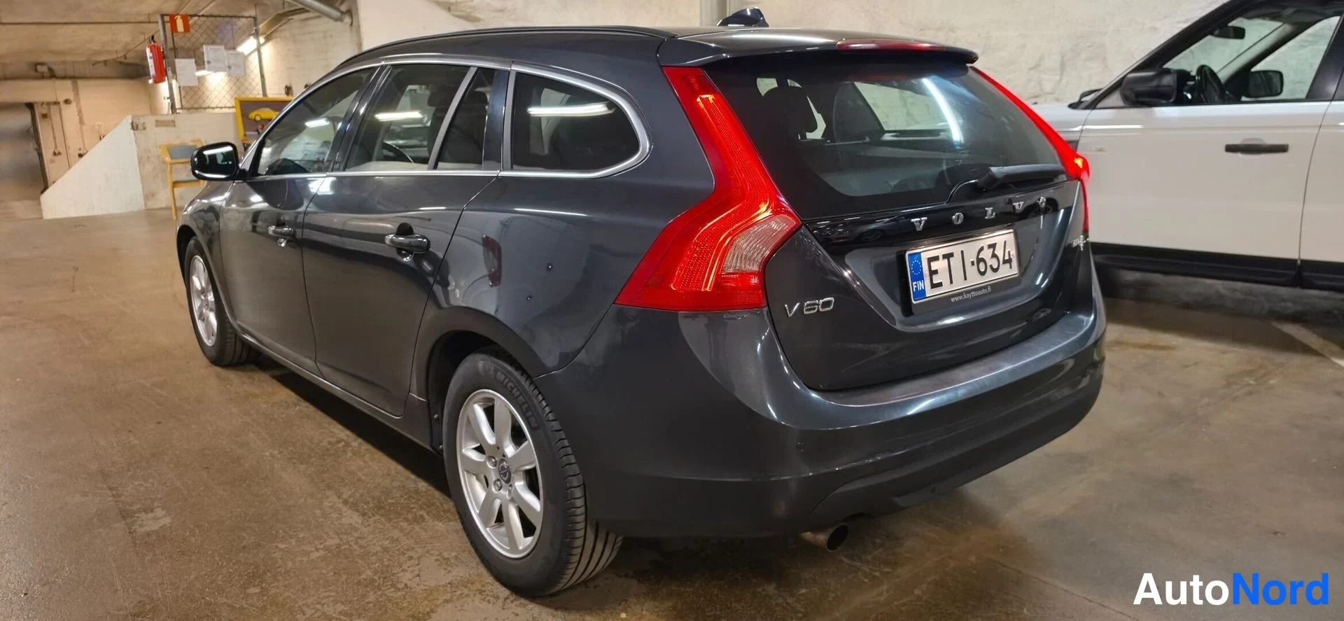 Takaviisto: Tummanharmaan farmariauton takakulma vasemmalta takaa kuvattuna autotallissa; näkyvissä takavalo, ruokakansi, V60-merkintä ja rekisterikilpi.
