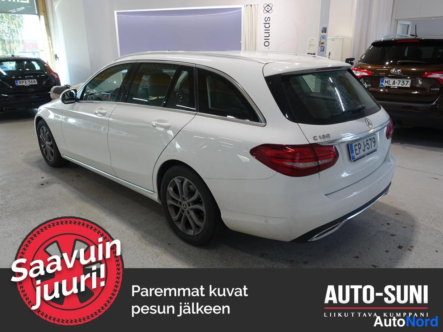 Takaviisto: Valkoinen Mercedes-Benz C-farmari takaosasta ja vasemmasta kyljestä kulmassa kuvattuna autosalissa, rekisteritunnus.