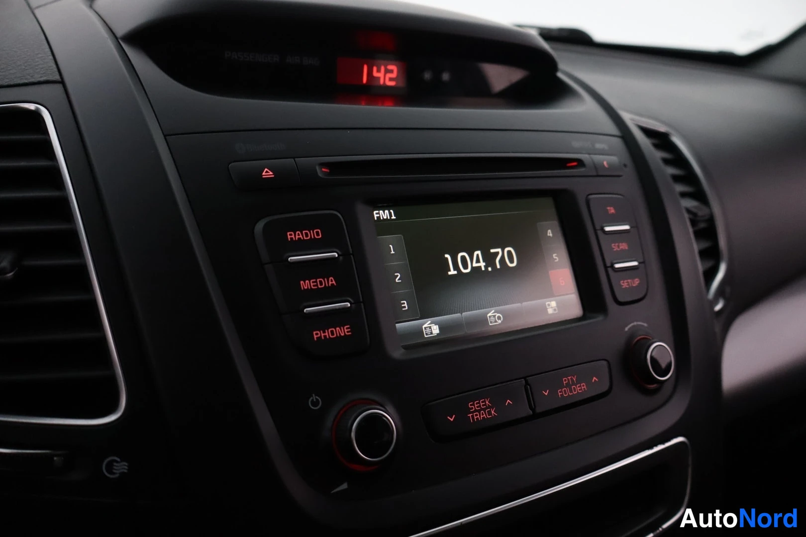 Yksityiskohta: Kojelaudan radio-/mediajärjestelmä lähikuvassa: RADIO-, MEDIA- ja PHONE-näppäimet, näytöllä FM1 104.70, ylhäällä kellon aika 1:42.