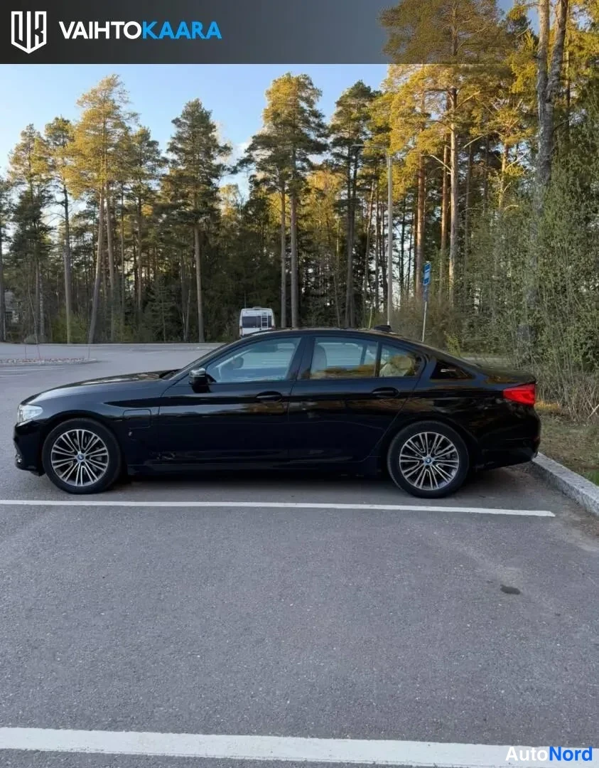 Sivulta: Musta BMW-sedan sivuprofiilista vasemmalle; kevytmetallivanteet, parkkipaikka metsän reunassa.