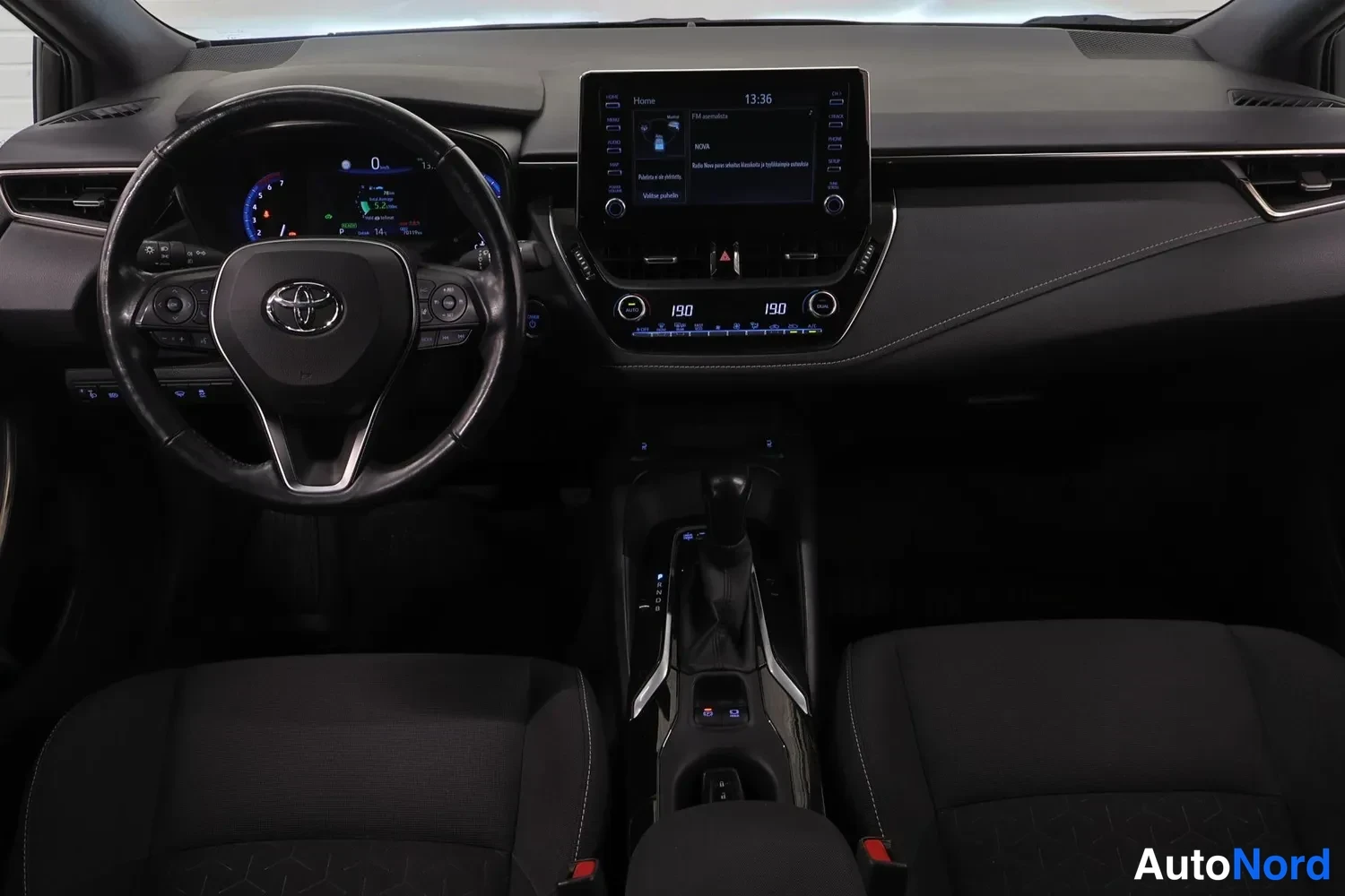 Muu: Ohjaamo suoraan edestä kuvattuna: Toyota-ohjauspyörä, mittaristo, suuri keskinäyttö, automaattivaihteiston valitsin ja kaksi etupenkkiä.
