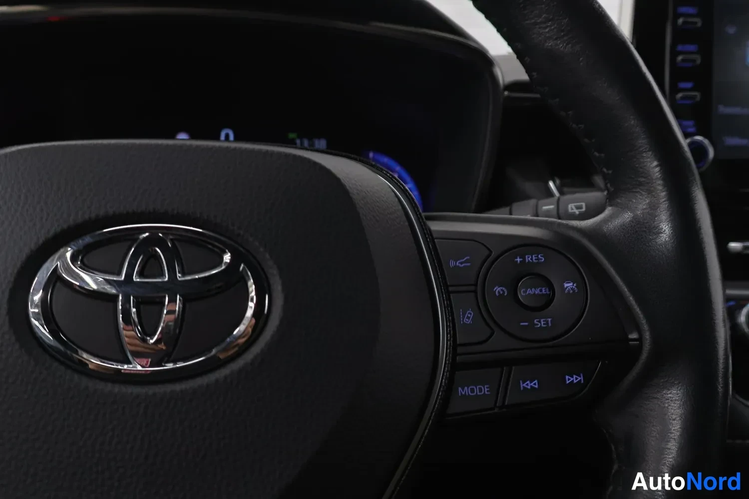 Yksityiskohta: Ohjauspyörän Toyota-merkki ja oikean puoleiset nappiryhmät: RES/CANCEL/SET (ruuvivakionopeudensäädin), MODE ja ääniohjausnappi.