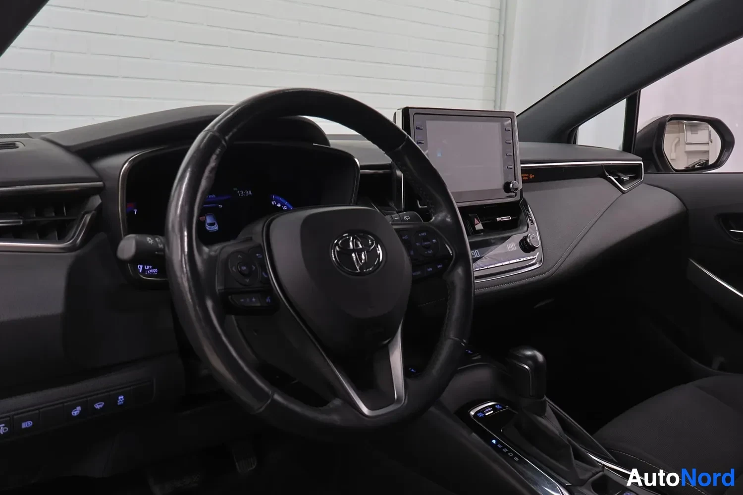 Muu: Ohjaamon näkymä vasemmalta: Toyota-merkkinen ohjauspyörä, digitaalinen mittaristo ja kosketusnäyttö keskitassulla.