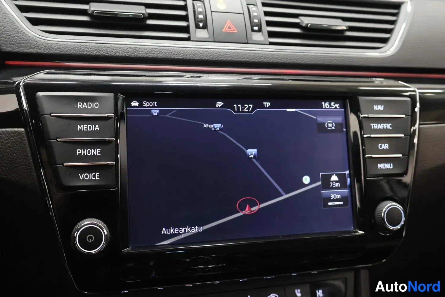 Yksityiskohta: Kojeiston keskikonsolin infotainment-näyttö, jolla näkyy navigointikartta merkinnöillä Aukeankatu ja Ahjo, kellonaika 11:27 ja lämpötila 16,5 °C. Näytön vasemma