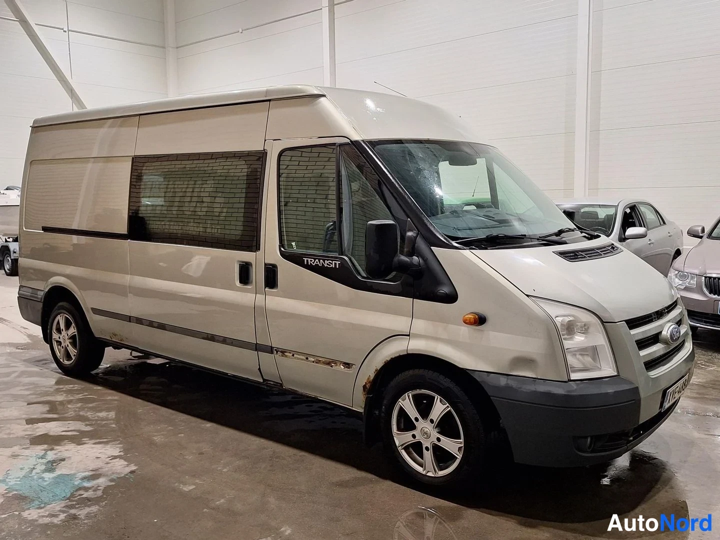 Etuviisto, nokka oikealle: Hopeanharmaa Ford Transit etuoikealta kolmessa neljäsosassa hallissa