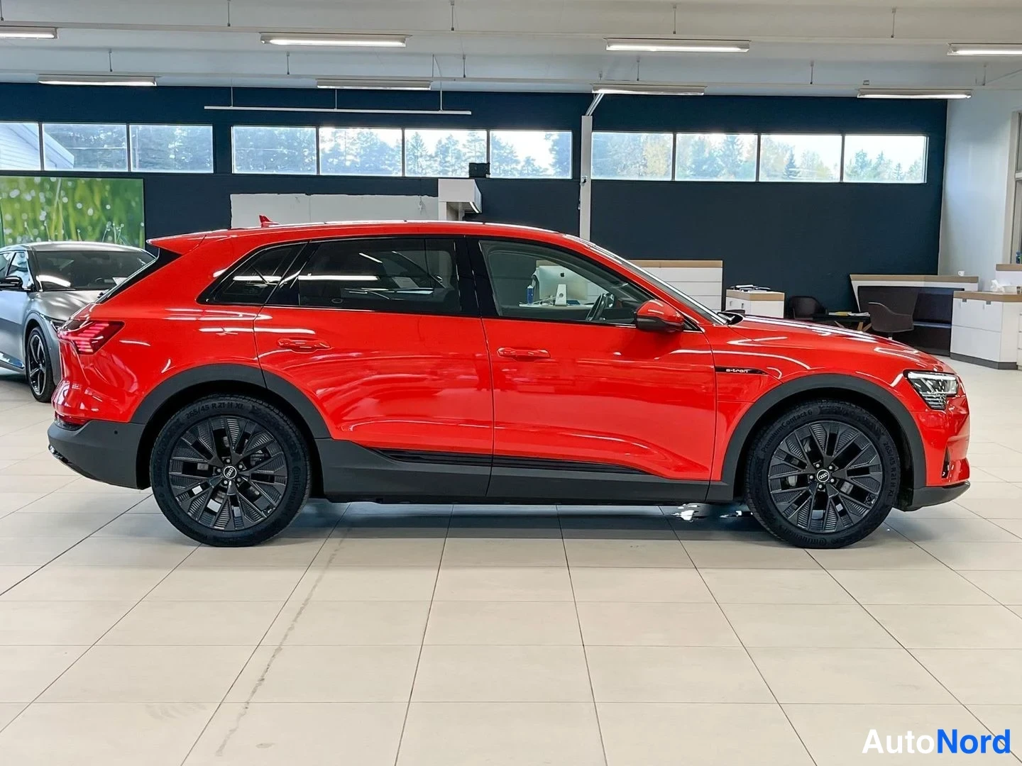 Etuviisto, nokka oikealle: Punainen Audi e-tron oikean kyljen sivunurkkakuvassa autosalissa; oikeat ovet, etu- ja takavalot sekä mustat vanteet näkyvissä.