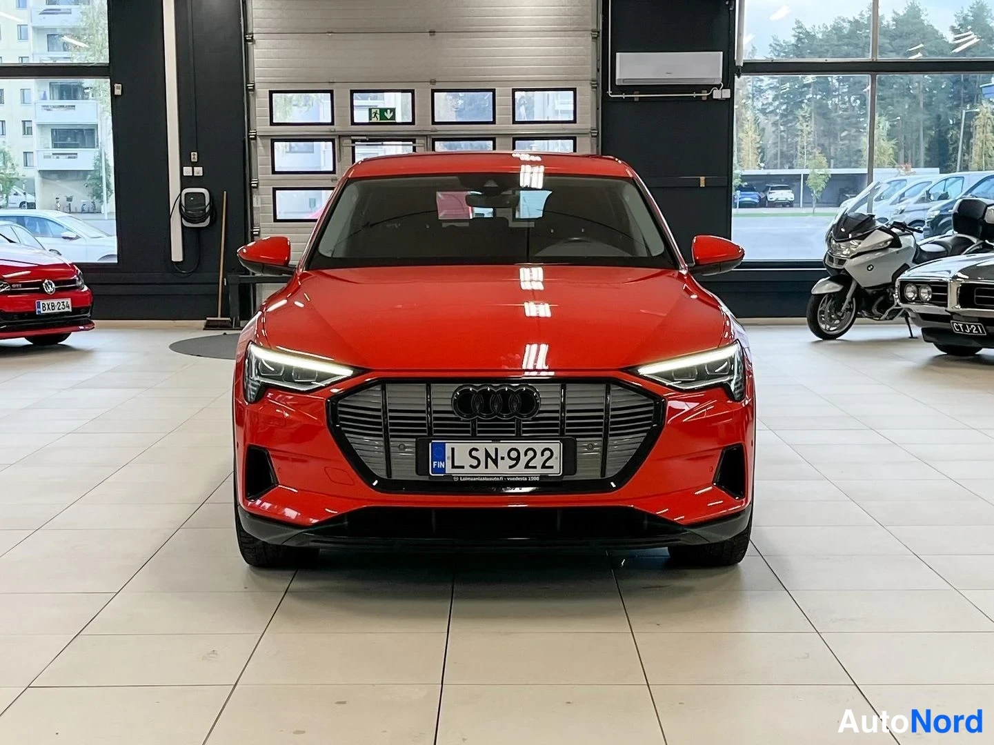 Edestä: Punaisen Audi e-tron -maasturin suora etukuva autosalissa; etugridi, Audi-merkki ja -rekisterikilpi näkyvät, LED-etuvalot ovat päällä.
