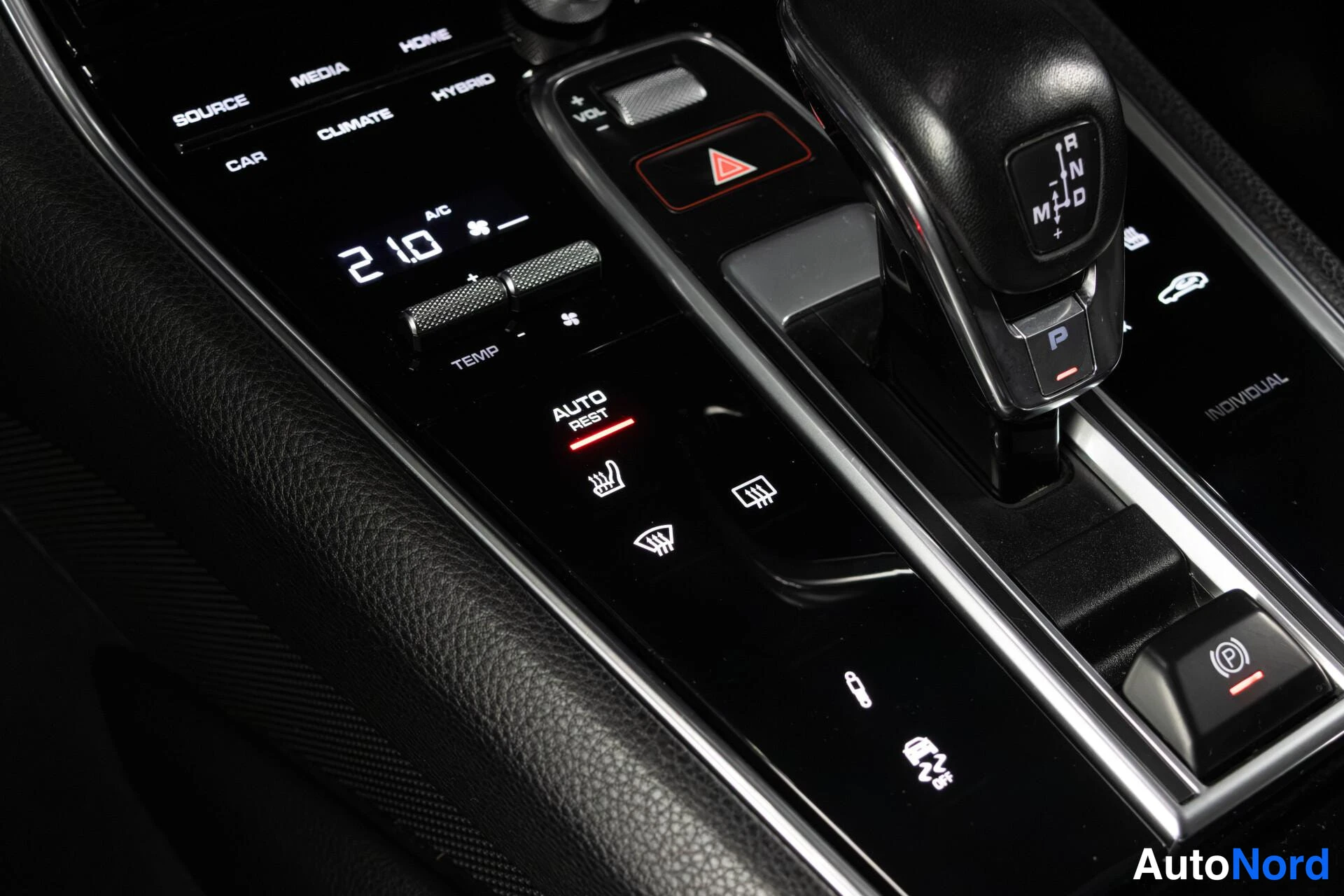 Yksityiskohta: Auton keskikonsolin lähikuva: automaattivaihteen valitsin (P R N D M), hybrid-merkintä, ilmastoinnin ohjaimet (TEMP, AC, AUTO REST), istuinlämmitys ja sähköinen