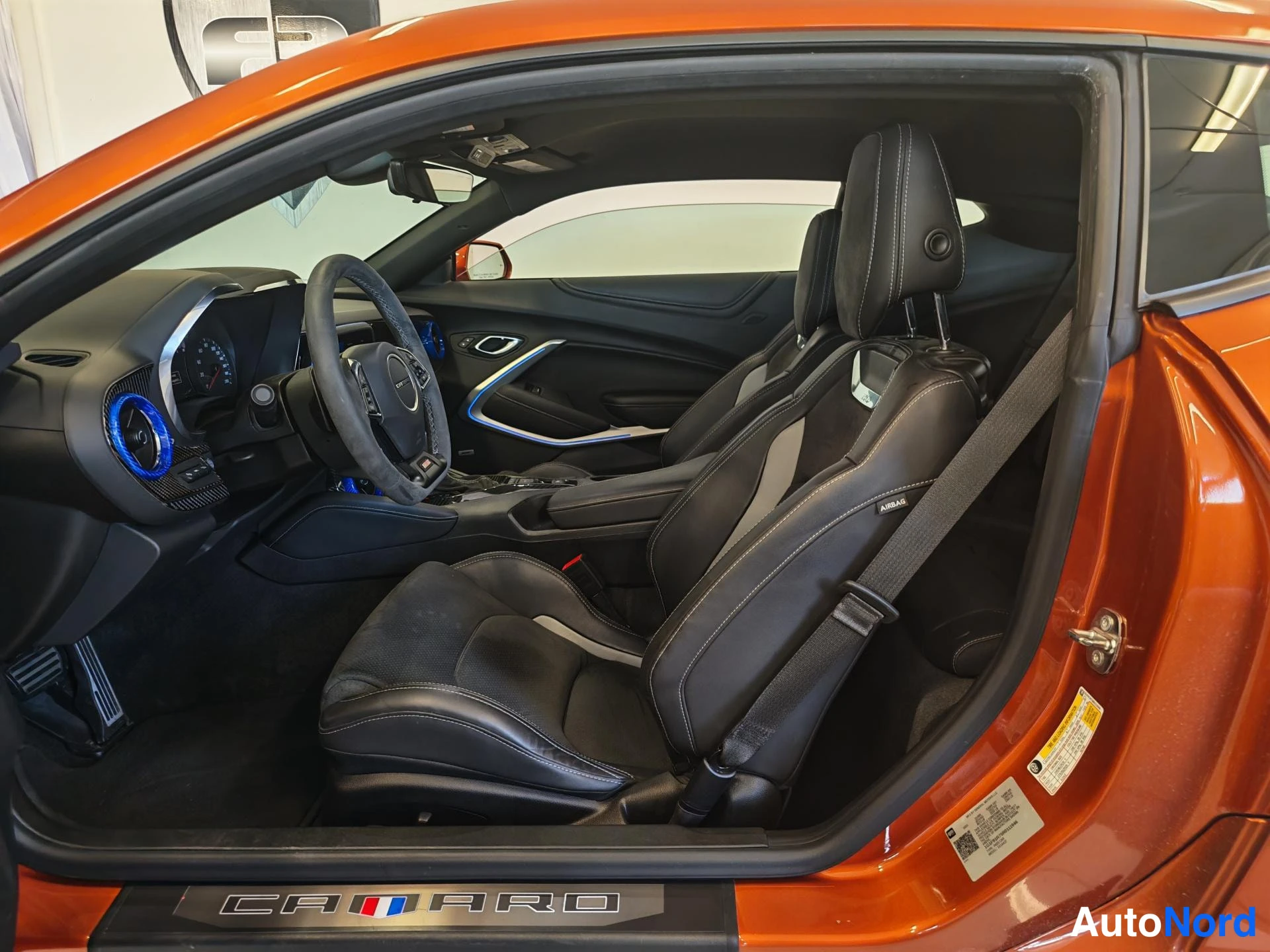 Sivulta: Oranssin coupé-mallisen auton avattu kuljettajanovi paljastaa mustan sisustan: alumiiniset polkimet, nahkaiset urheiluistuimet ja Camaro-logolla varustettu kynn