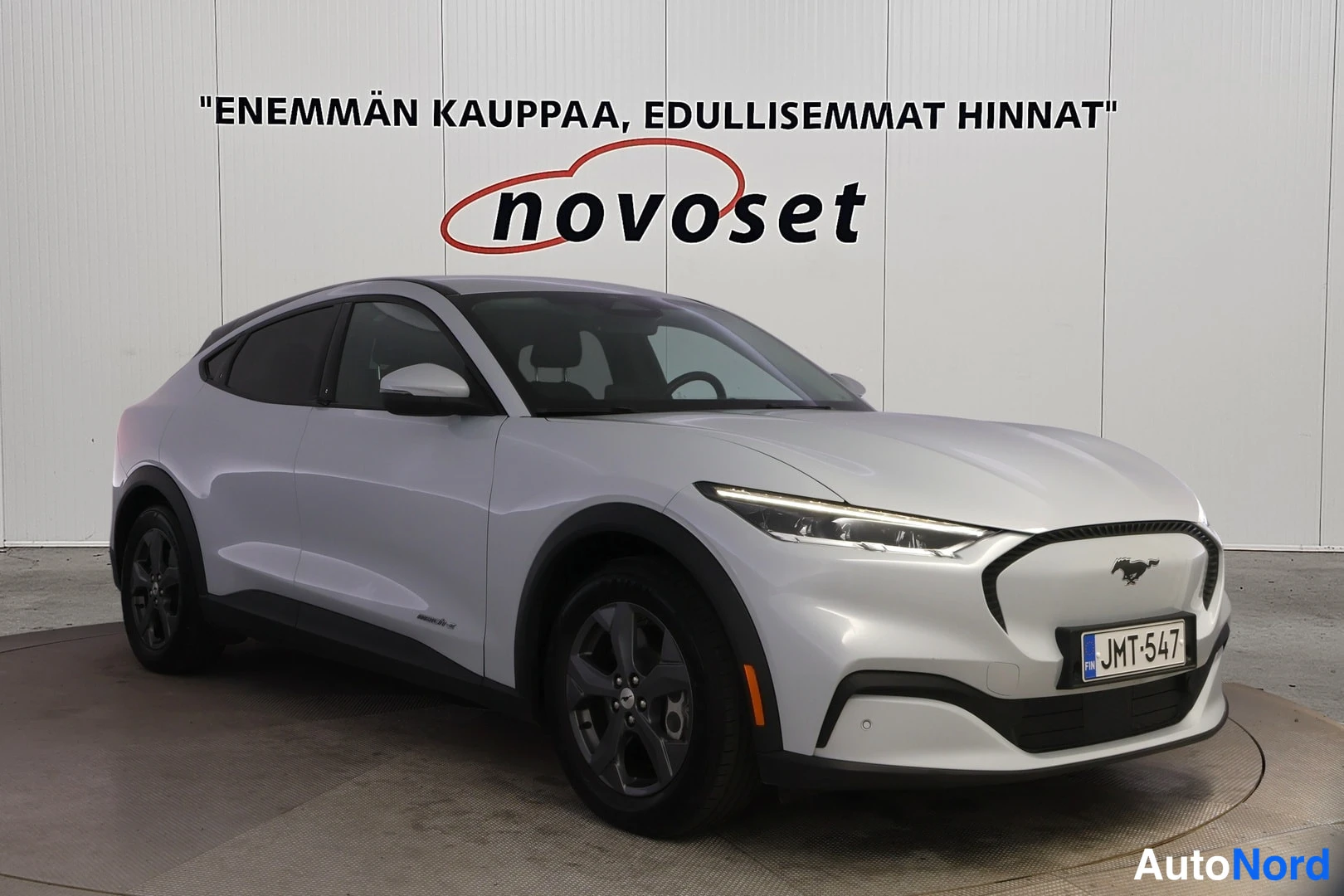 Etuviisto, nokka oikealle: Hopeanvärinen Ford Mustang Mach-E etuviistosta vasemmalta, rekisteritunnus, novoset-mainosseinä taustalla.