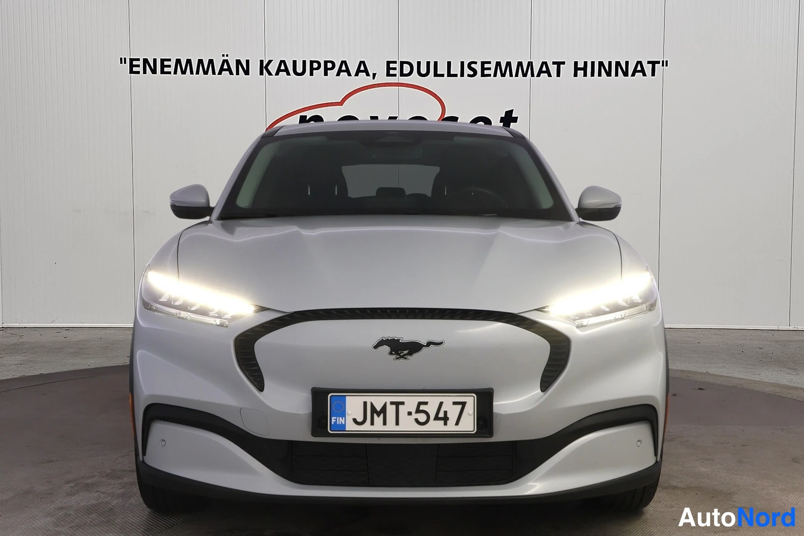 Edestä: Ford Mustang Mach-E suoraan edestäpäin, ajovalot päällä, rekisteritunnus, hopeanvärinen kori.