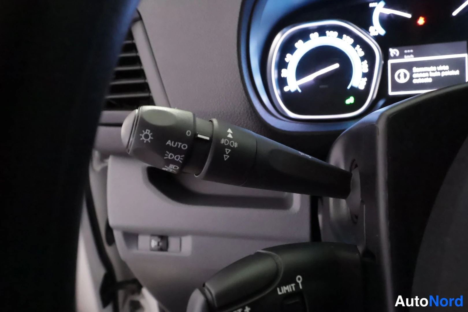 Yksityiskohta: Sisätilakuvat: kevytvipu, jossa on merkinnät AUTO, DO ja sumunestopainikkeet; kojelautanäyttö tummassa.