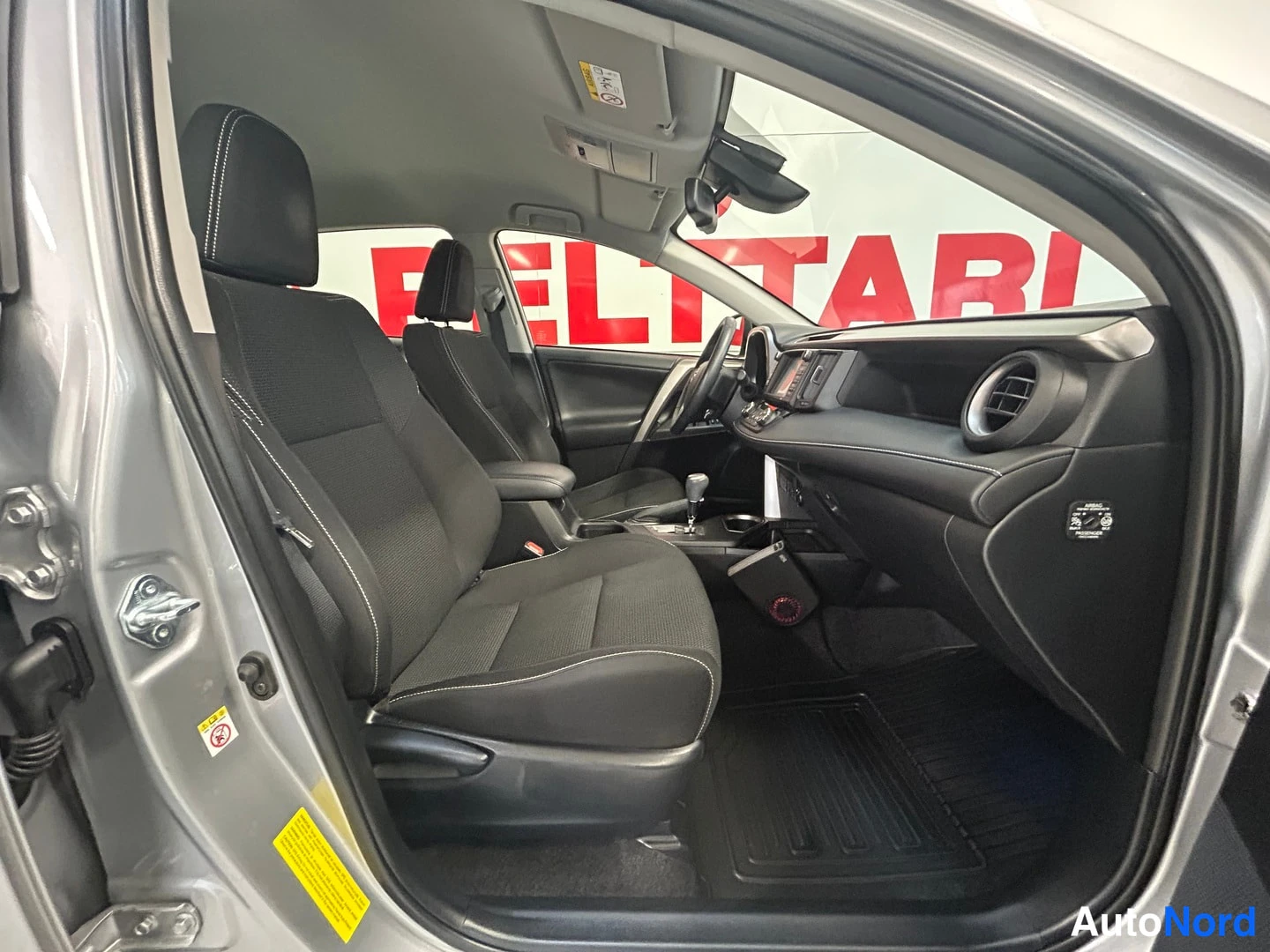 Muu: Sisäkuva Toyota RAV4:stä matkustajan puolen oven kautta: etuistuimet mustaa kangasta, kojelauta ja keskikonsoli näkyvät.