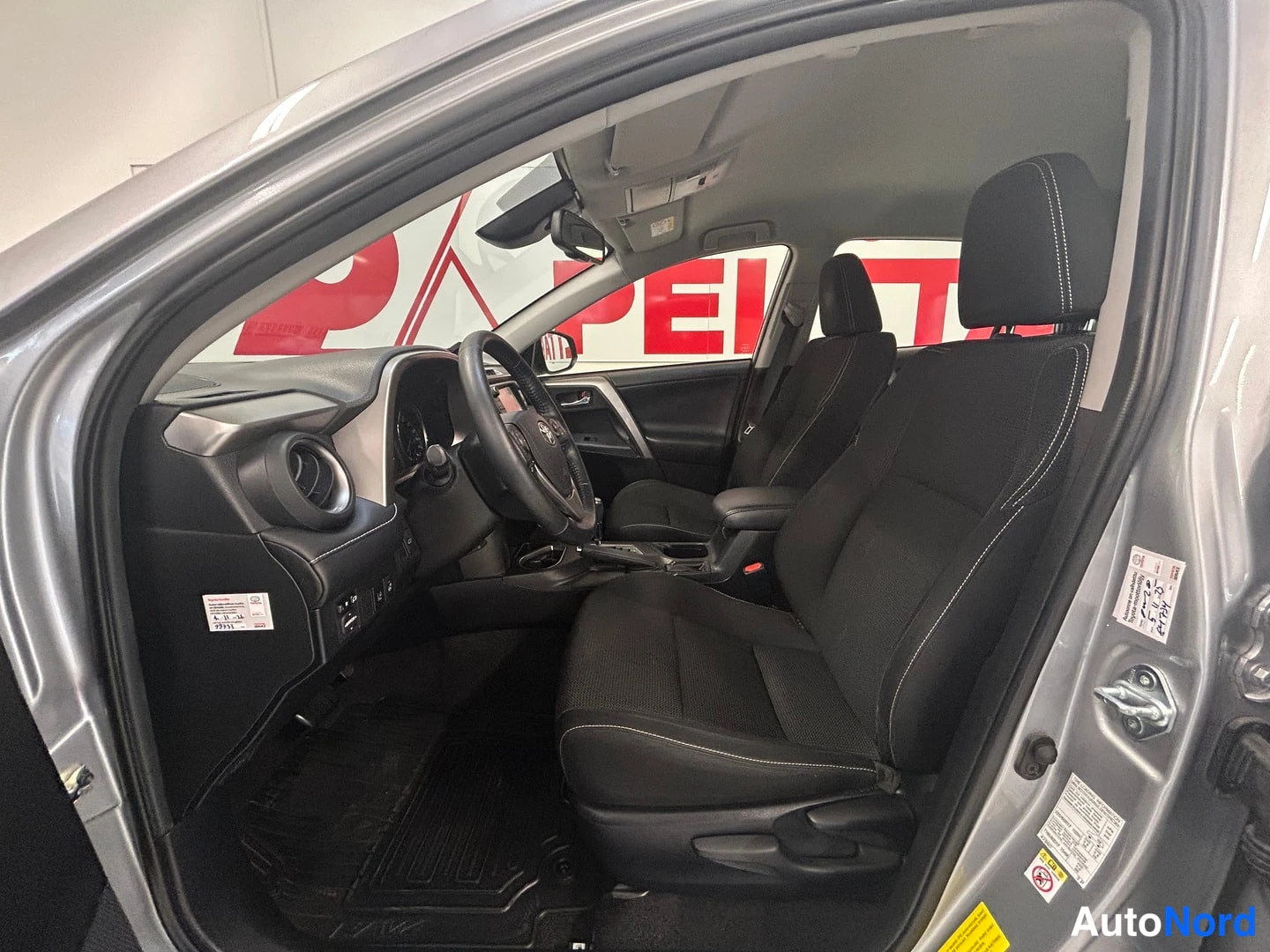 Muu: Sisäkuva Toyota RAV4:stä: avattu kuljettajan ovi, ohjaamo ja kaksi etuistuinta mustalla kangasverhoilulla, kojelauta ja ohjauspyörä näkyvät.
