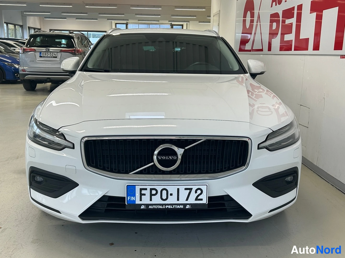 Edestä: Valkoisen farmariauton etunäkymä sisätiloissa, VOLVO-teksti etusäleikössä, rekisterikilpi FP0-172.