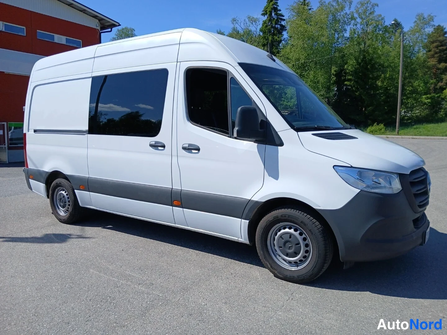 Etuviisto, nokka oikealle: Mercedes-Benz Sprinter -pakettiauto valkoisena kylki ja keula näkyvissä oikealta kuvakulmasta, tummat sivuikkunat ja punatiilinen rakennus taustalla.