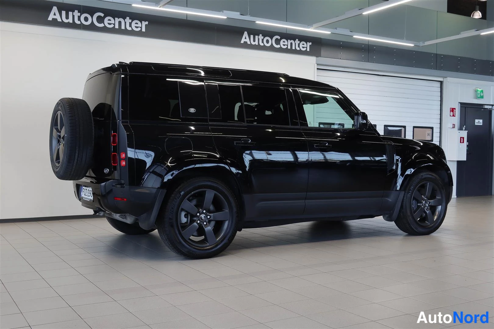 Takaviisto: Musta Land Rover Defender takaviistosta sisätiloissa; takana ulkoinen varapyörä, punaiset takavalot ja tummat vanteet.