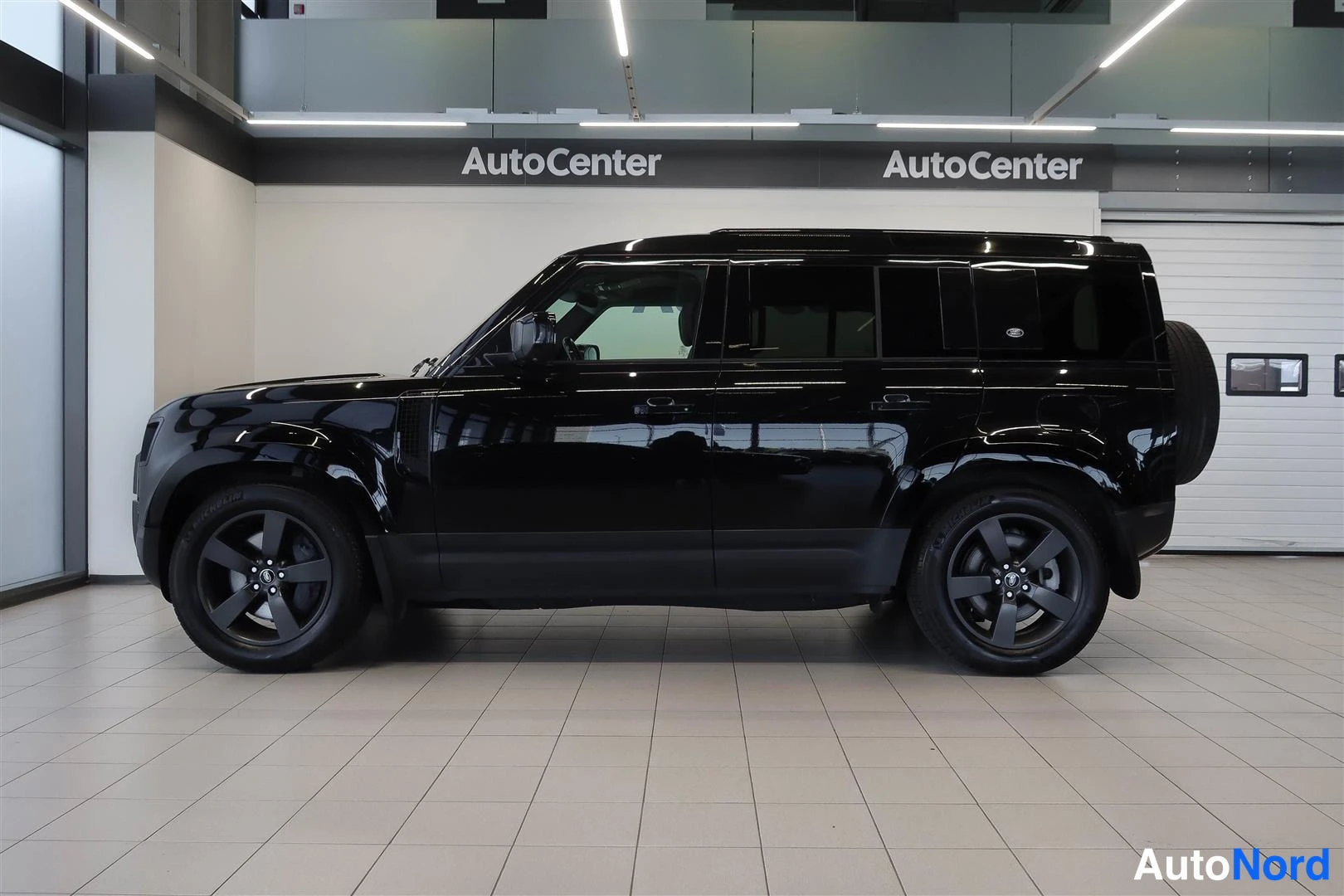 Sivulta: Musta Land Rover Defender sivulta sisätiloissa; sileä kylkipinta, tummat vanteet ja takaosan varapyörän kiinnike näkyy osittain.