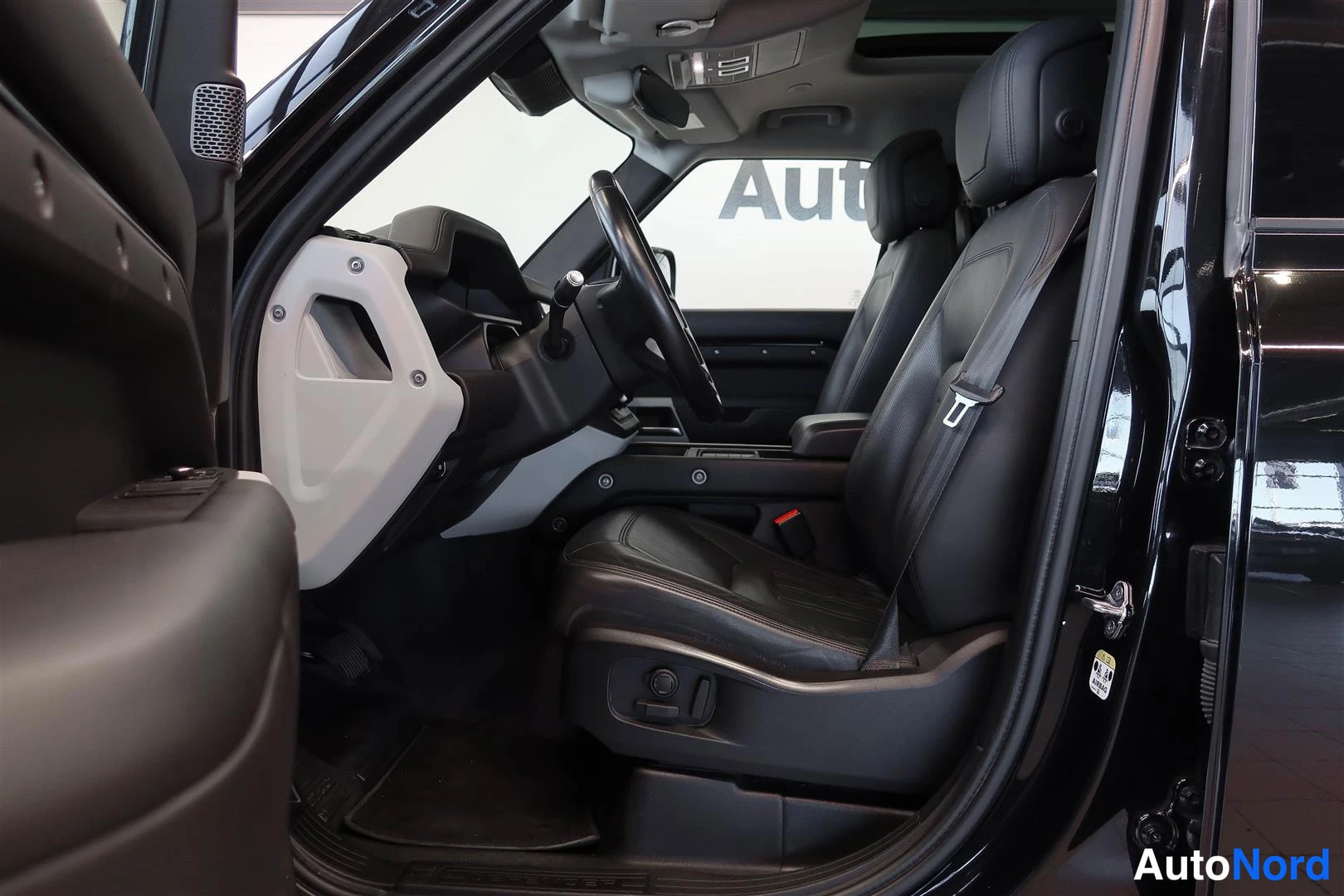 Muu: Land Rover Defenderin etupenkki ja ohjaamo vasemmalta ovenraosta kuvattuna: mustat nahkapenkit, kevytkatteet ja osa kojelautaa.