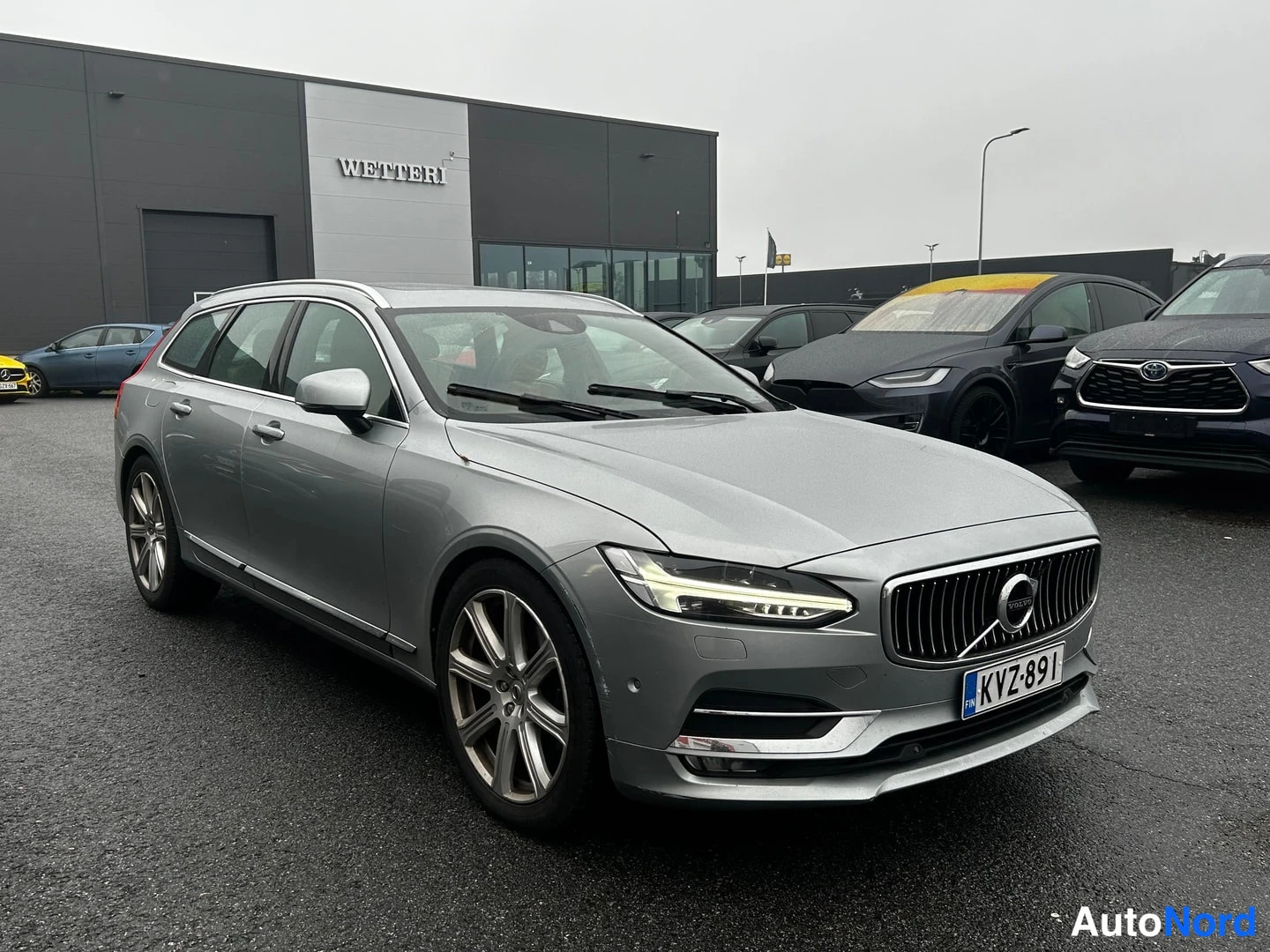 Etuviisto, nokka oikealle: Etukulma hopeanharmaasta Volvo V90 -farmarista parkkipaikalla, nokka oikealle, rekisterikilpi.