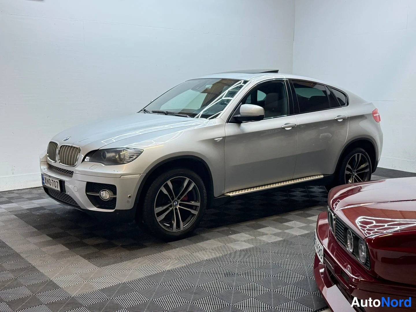 Etuviisto, nokka vasemmalle: BMW X6 etukulmasta, neontelalla, etupyörän ja kyljen näkyvä runko sisätiloissa.