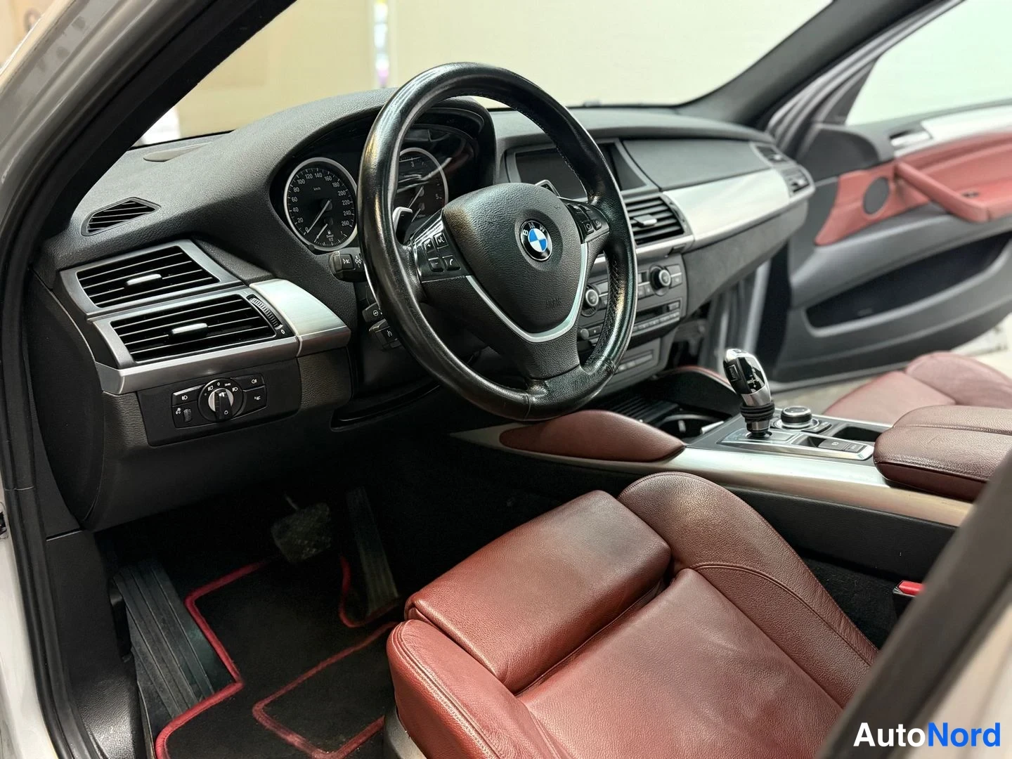 Muu: Kojelaudan ja etuistuinten tarkempi kuva: BMW-ohjauspyörä, mittaristo, keskikonsolin vaihtaja ja punaruskea nahkaverhoilu.