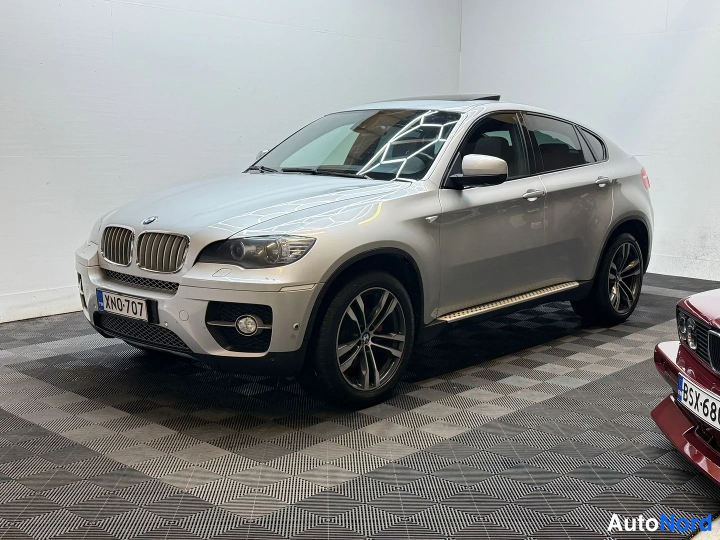 Etuviisto, nokka vasemmalle: BMW X6 etukulmasta kuvattuna, -kilpi, neontelalla, toinen auto vieressä.