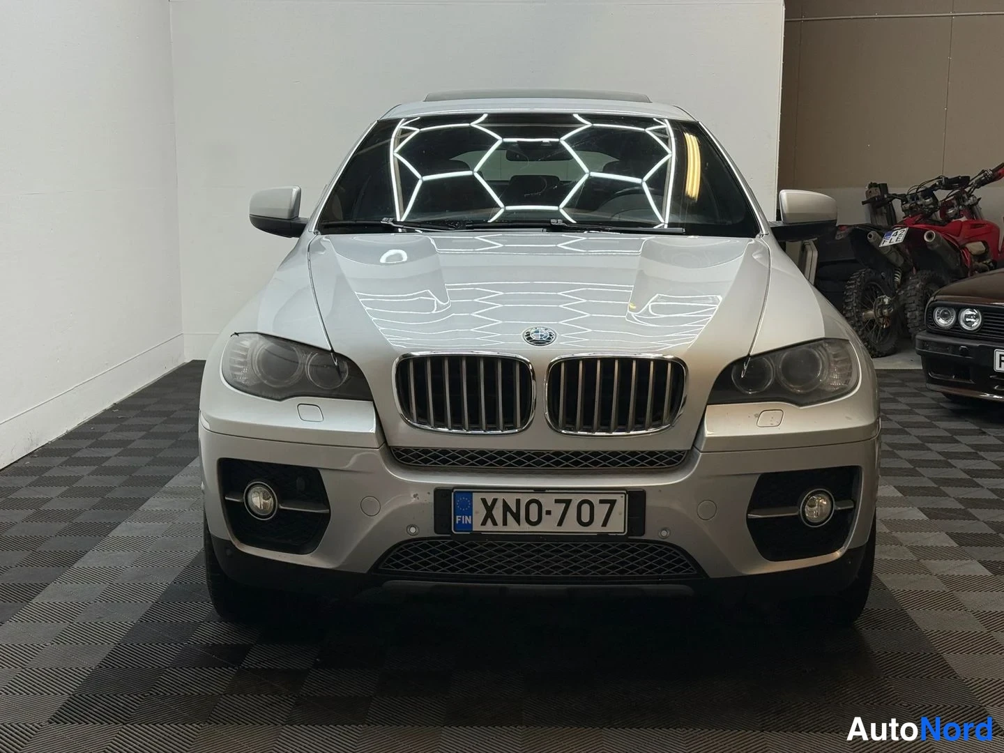 Edestä: BMW X6:n etupää kohtisuoraan kuvattuna valoisaan autotalliin, rekisteritunnus, hopeanvärinen kori.