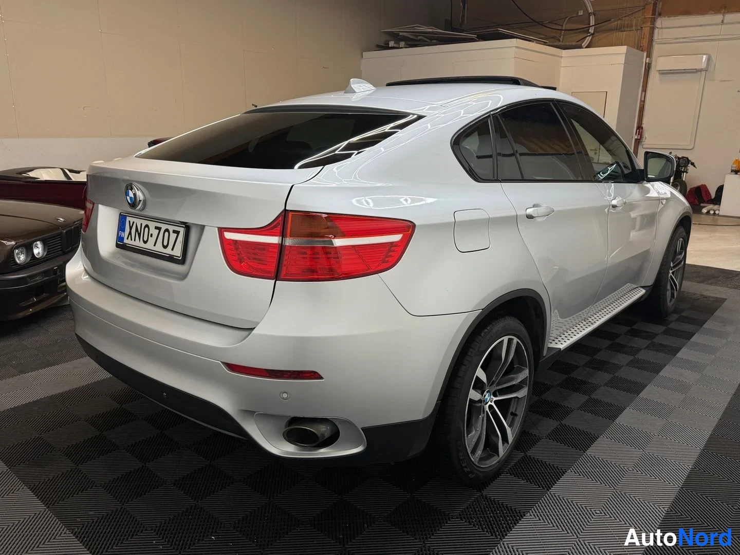 Takaviisto: Hopeanvärinen BMW:n takapää ja sivuprofiili sisätiloissa, rekisterikilpi, panoramic-katto, takavalo ja pakanpput kiinnittäneet huomiota.