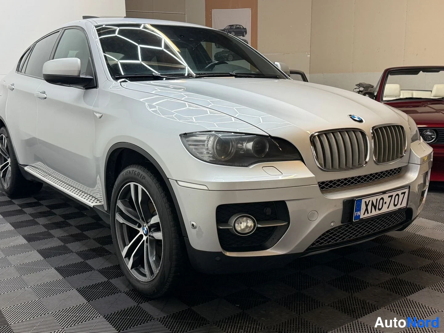 Etuviisto, nokka oikealle: Hopeanvärinen BMW X6 etukulmasta oikealta, rekisterikilpi, kuvattu sisätilassa mustavalkoisella lattialla.