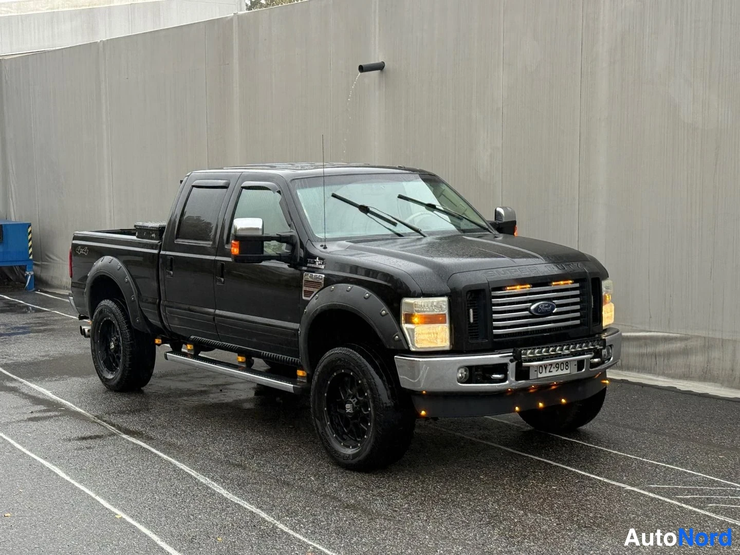 Etuviisto, nokka oikealle: Musta Ford F-250 lava-auto etuviistosta valossa, jatketut lokasuojat ja lisävalot, rekisterikilpi, märkä parkkipaikka harmaalla seinällä.