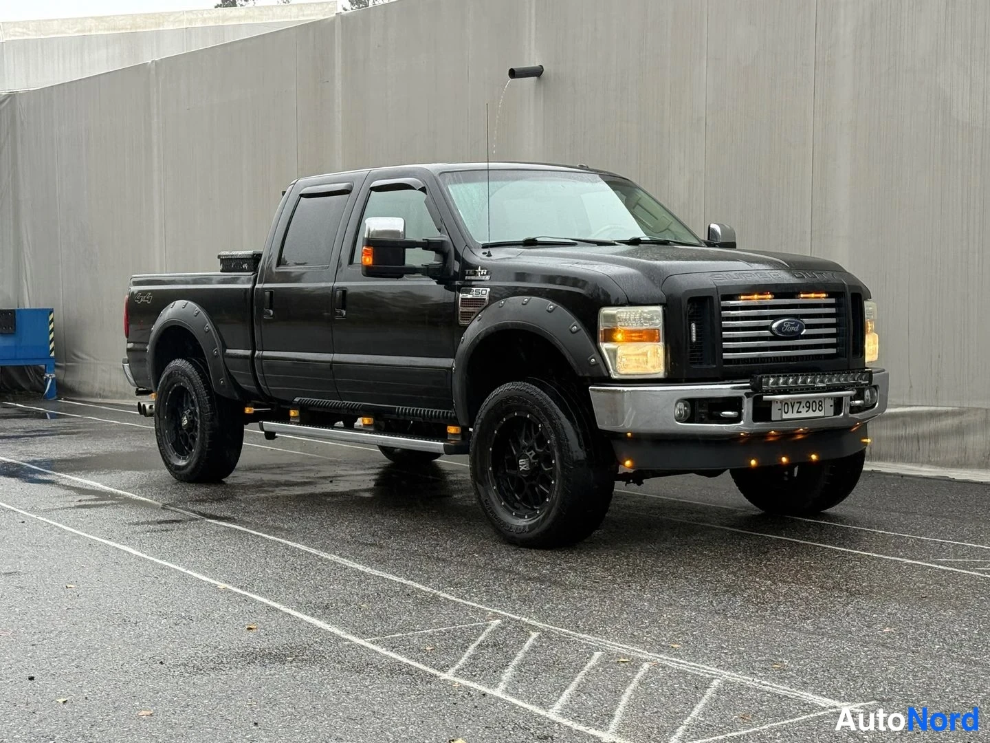 Etuviisto, nokka oikealle: Musta Ford F-250 lava-auto etuviistosta, mustat vanteet, kromipuskurit ja LED-palkki etupuskurissa, märkä asfaltti.