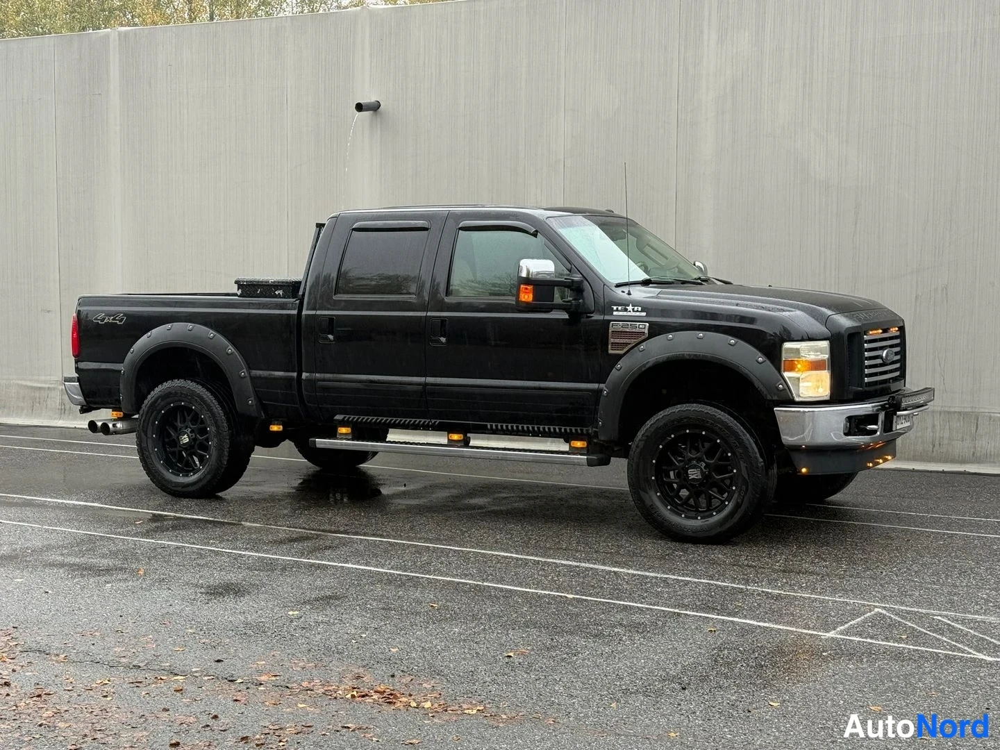 Etuviisto, nokka oikealle: Musta Ford F-250 lava-auto etuviistosta, jalustat ovien alla ja 4x4-merkintä lavan sivulla, märkä parkkipaikka.