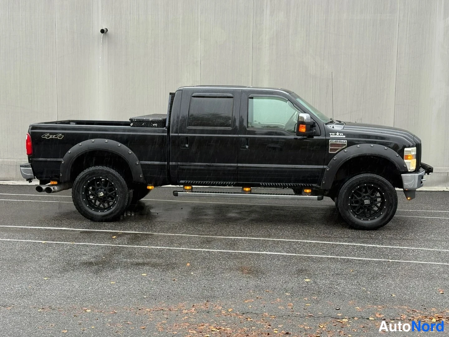 Etuviisto, nokka oikealle: Musta Ford F-250 lava-auto sivusta etuviistoon, sadekeli, mustat vanteet ja jalustat, takapyhreet sumussa.