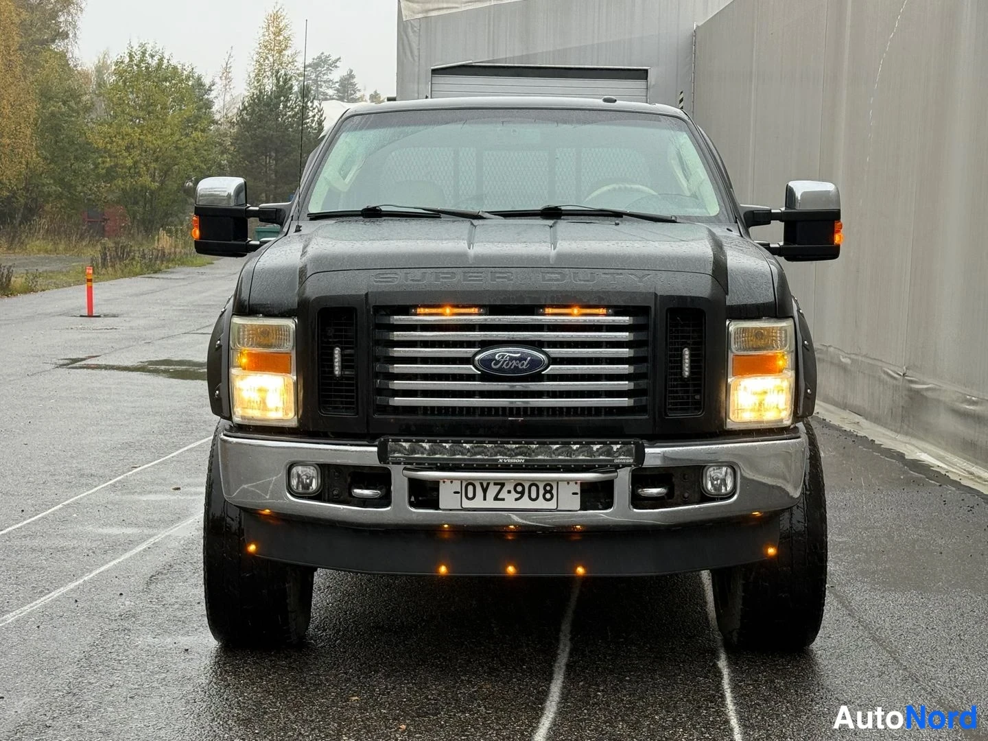 Edestä: Mustan Ford Super Dutyn etunäkösuora kuva; rekisterikilpi, etutulet päällä, Ford-logo säleikössä, lisävalo pyörätuolitelineessä ja LED-palkki puskurissa.