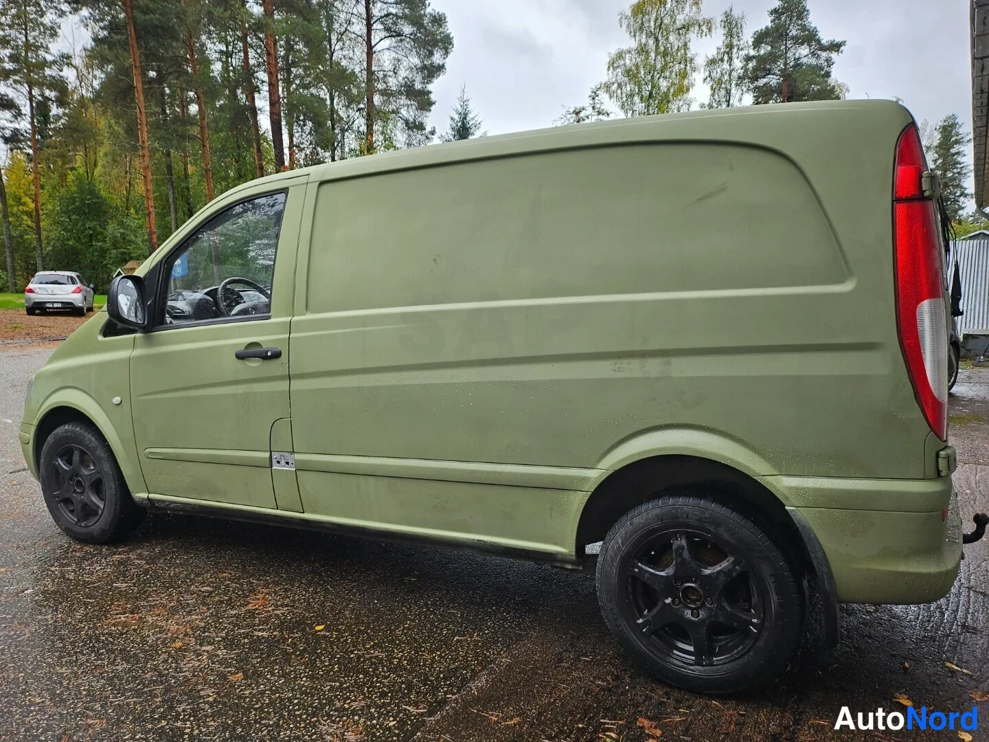 Takaviisto: Vihertävän ruskeanvärinen Mercedes-Benz Vito -pakettiauto kuvattu takaviistosta; sivut ja takavalot näkyvät, mustat peltivanteet, märkä asfaltti ja metsä tausta
